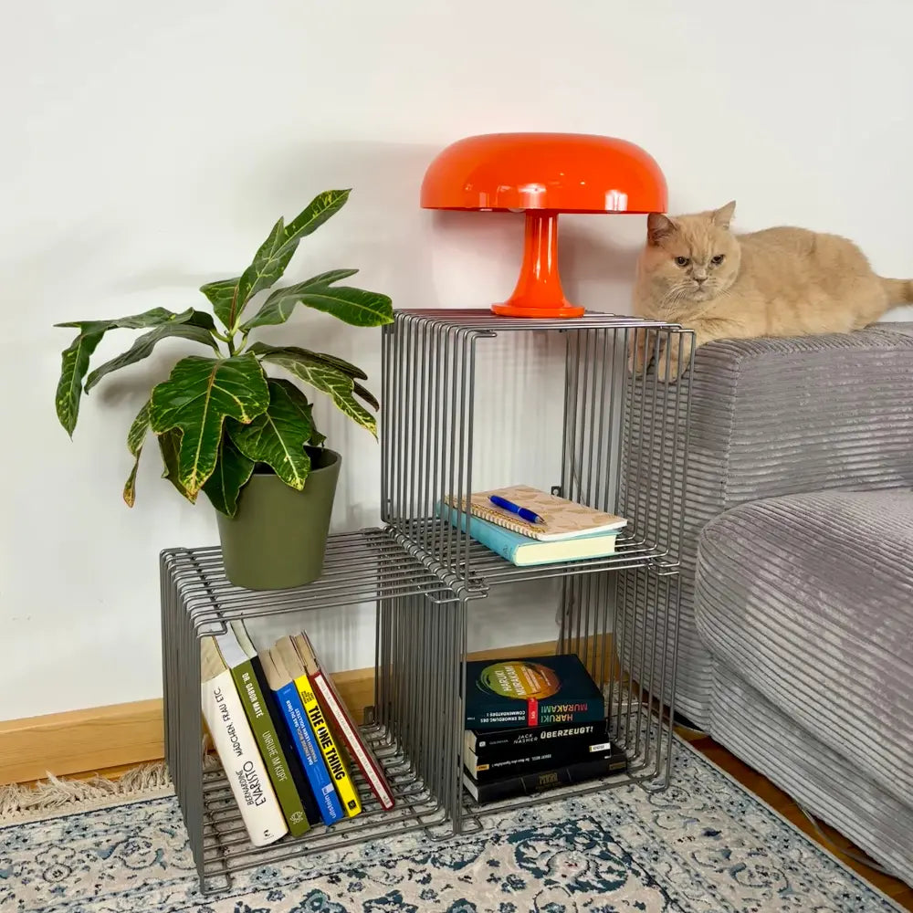 Wire side table with books, a plant, and an orange lamp next to a gray sofa with a cat.