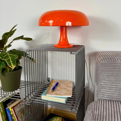 Orange lamp on a metal shelf with books, a plant, and a notebook.